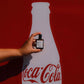 Hand holding a small bottle in front of a large Coca-Cola bottle silhouette on a red background
