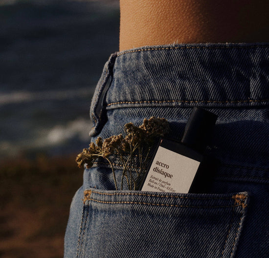 An image of a bottle of accrodisiaque perfume in a blue jean pocket with a faded background of a beach. In the pocket there is also a few brown/pink flowers. 