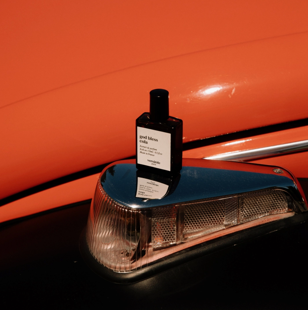 A brown glass bottle of Versatile Paris God Bless Cola perfume sits on a metallic object, in front of an orangey-red metallic curved background, which looks like it might be a car but we can't tell 