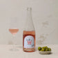 A clear glass bottle with a pale blue label with pink writing stands next to a glass of rosé wine and a bowl of olives