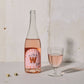 Bottle of Wednesday’s Domaine 0.0% Elan Alcohol-Free Rosé Wine with a pink hue, embodying refreshing acidity and summer fruit notes. It stands next to a glass of rosé wine and 2 dice