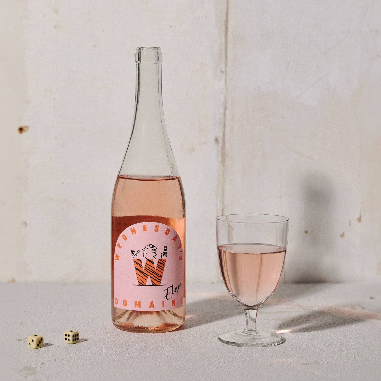 Bottle of Wednesday’s Domaine 0.0% Elan Alcohol-Free Rosé Wine with a pink hue, embodying refreshing acidity and summer fruit notes. It stands next to a glass of rosé wine and 2 dice