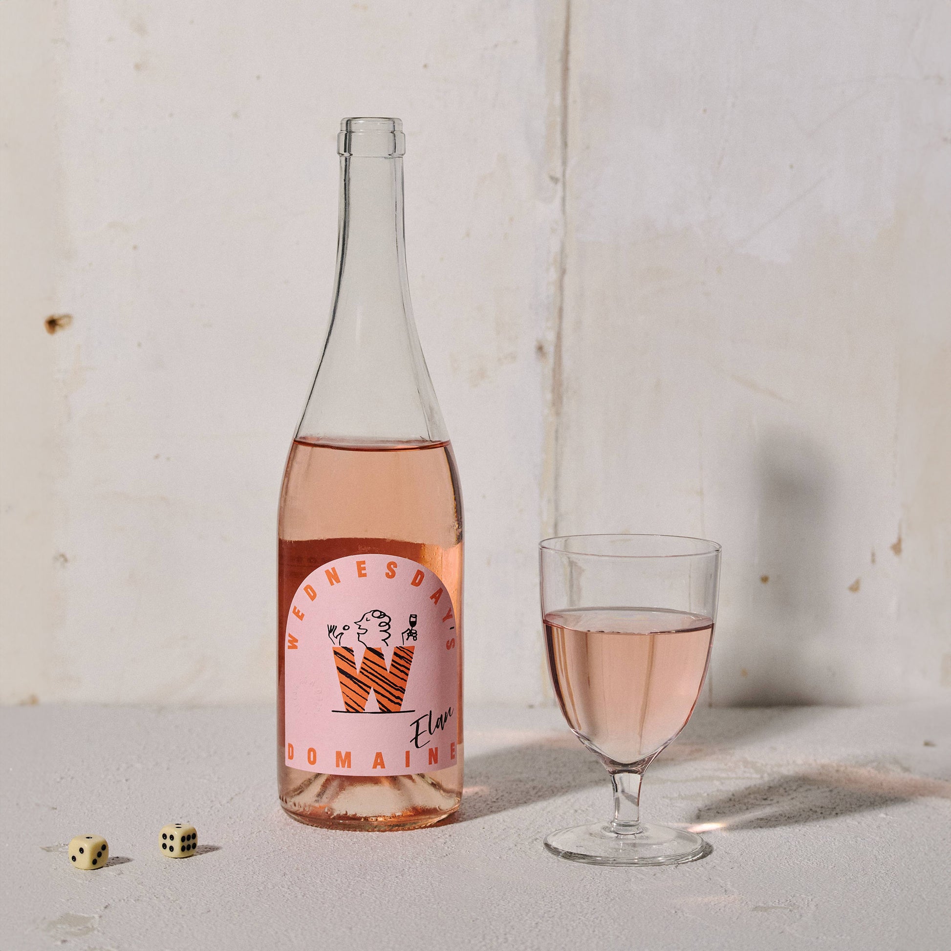 Bottle of Wednesday’s Domaine 0.0% Elan Alcohol-Free Rosé Wine with a pink hue, embodying refreshing acidity and summer fruit notes. It stands next to a glass of rosé wine and 2 dice