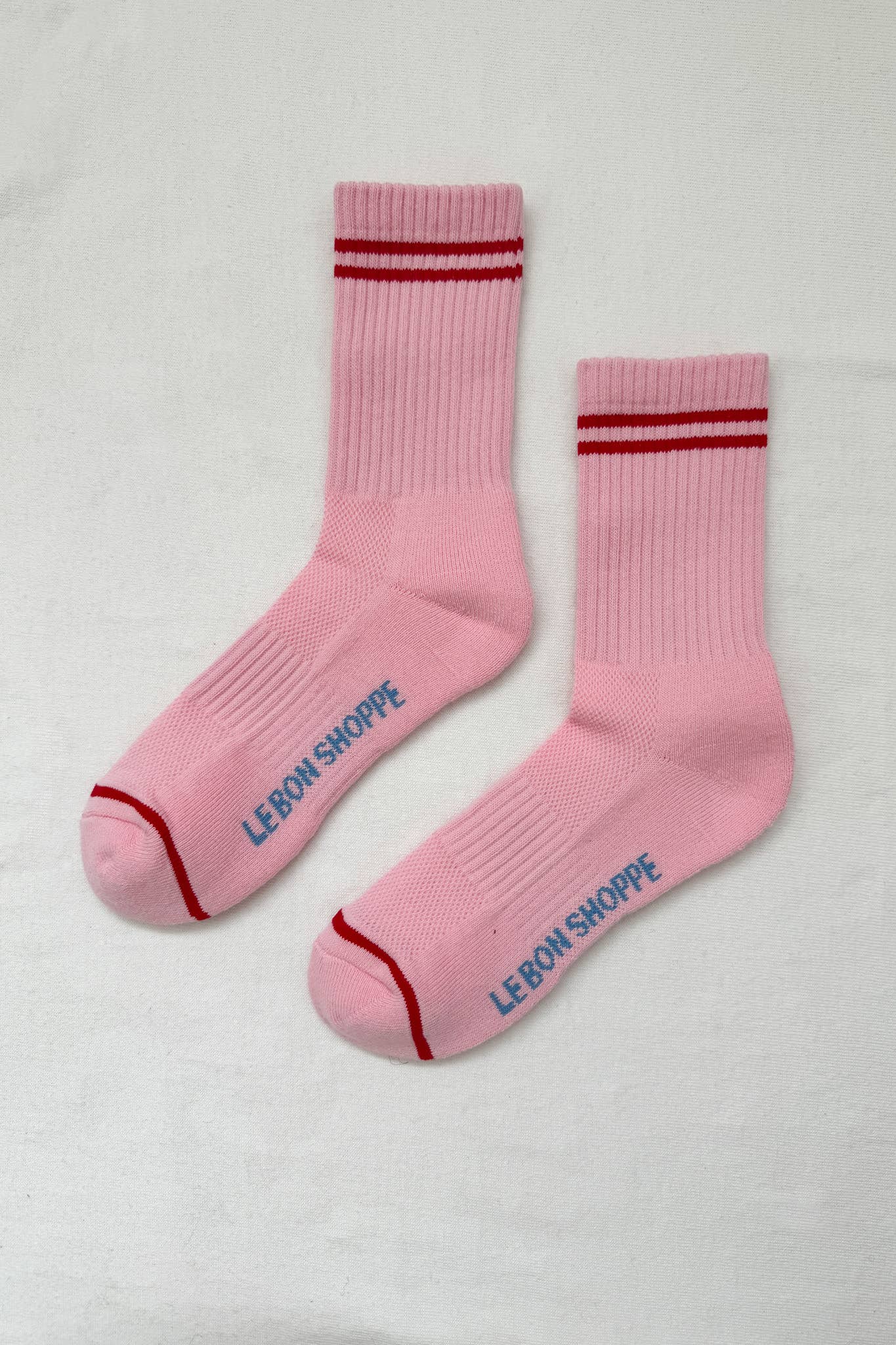 A pair of Le bon shoppe amour pink boyfriend socks which are pink in colour with red stripes at the top and near the toes.It says Le Bon shoppe on the sole of the socks in blue colour. The socks are kept against a white background. 