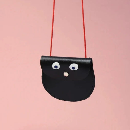 An oval shaped black small pocket purse with red thin rope as the bag strap hangs against a pink background. Printed onto the bag are two white googly eyes with black pupils and a small silver press button which acts as the nose and opens/closes the purse. 