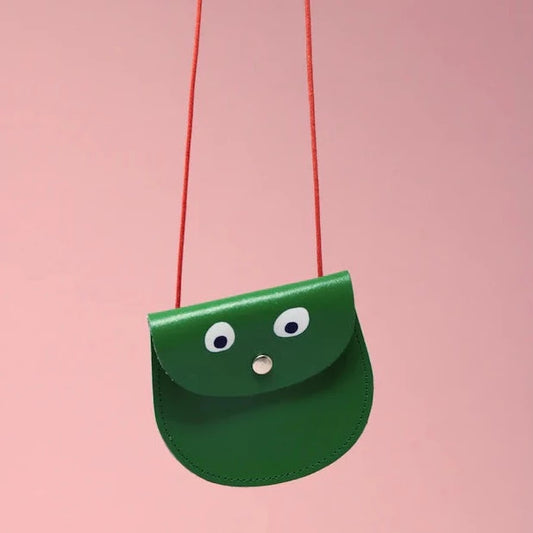 An oval shaped bright green small pocket purse with red thin rope as the bag strap hangs against a pink background. Printed onto the bag are two white googly eyes with black pupils and a small silver press button which acts as the nose and opens/closes the purse. 