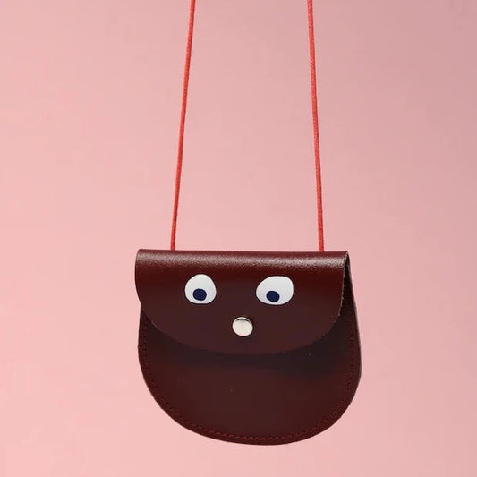 An oval shaped burgundy small pocket purse with red thin rope as the bag strap hangs against a pink background. Printed onto the bag are two white googly eyes with black pupils and a small silver press button which acts as the nose and opens/closes the purse. 