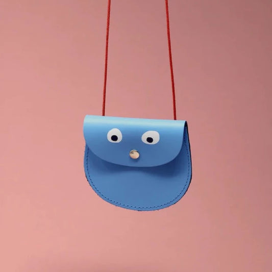 An oval shaped light blue small pocket purse with red thin rope as the bag strap hangs against a pink background. Printed onto the bag are two white googly eyes with black pupils and a small silver press button which acts as the nose and opens/closes the purse.  