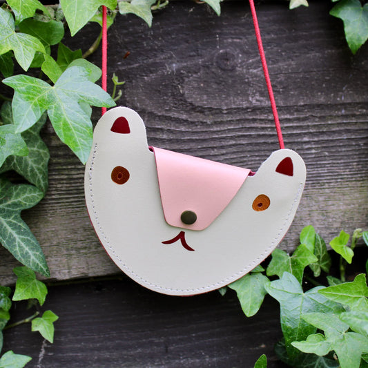 A cream pocket purse in the shape of a bears head hanging in front of what appears to be a wooden fence with leaves surrounding it. The flap of the bag is a pale pink that acts as the nose of the bear and has a a black button. The eyes have a red/orange shade and the ears are crimson. Beneath the flap the mouth is also crimson. The strap of the bag is thin red rope . 