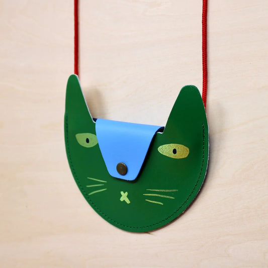 A dark green pocket purse in the shape of a cats head hanging in front of a cream wooden background. The flap of the bag is a light blue that acts as the nose of the cat and has a a black button. The eyes are oval shaped and metallic gold with black pupils. Beneath the flap, there are 3 metallic  gold whiskers on either side with a metallic gold cross in the middle, acting as a mouth.The strap of the bag is thin red rope. 