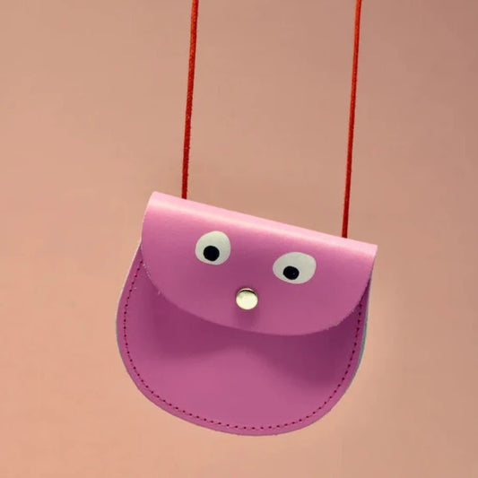 An oval shaped hot pink small pocket purse with red thin rope as the bag strap hangs against a pink background. Printed onto the bag are two white googly eyes with black pupils and a small silver press button which acts as the nose and opens/closes the purse. 