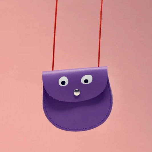 An oval shaped lilac small pocket purse with red thin rope as the bag strap hangs against a pink background. Printed onto the bag are two white googly eyes with black pupils and a small silver press button which acts as the nose and opens/closes the purse. 