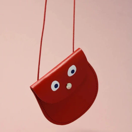 An oval shaped red small pocket purse with red thin rope as the bag strap hangs against a pink background. Printed onto the bag are two white googly eyes with black pupils and a small silver press button which acts as the nose and opens/closes the purse.