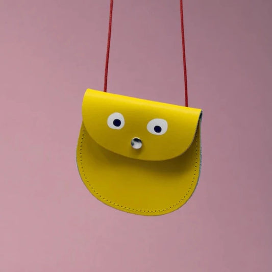 An oval shaped yellow small pocket purse with red thin rope as the bag strap hangs against a pink background. Printed onto the bag are two white googly eyes with black pupils and a small silver press button which acts as the nose and opens/closes the purse. 