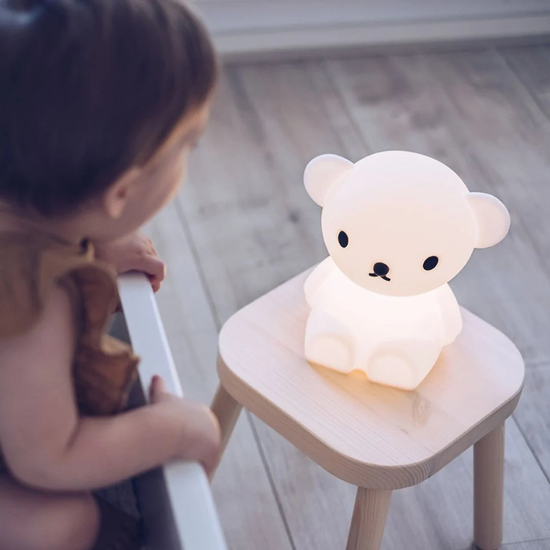 A young child sitting in a crib looking at a glowing Boris First Light Lamp placed on a wooden stool in a room.