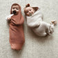 Two babies wrapped in orange coloured and off white coloured wool cocoons lying on a beige background