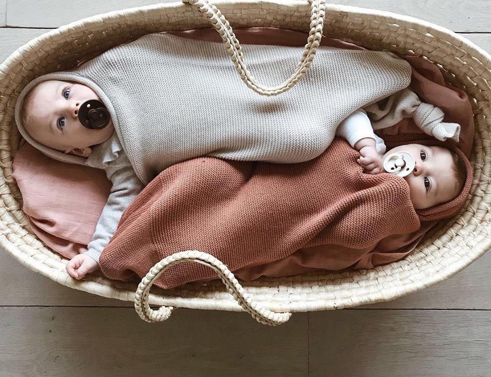 Two babies in an orange and off white wool cocoons placed in a rope basket on a wooden panel floor.