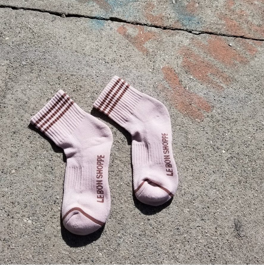 A pair of Bellini pink ribbed crew socks with brown stripe details, laid flat on a textured concrete surface with faint graffiti, highlighting the Le Bon Shoppe branding on the sole and contrast toe accents.