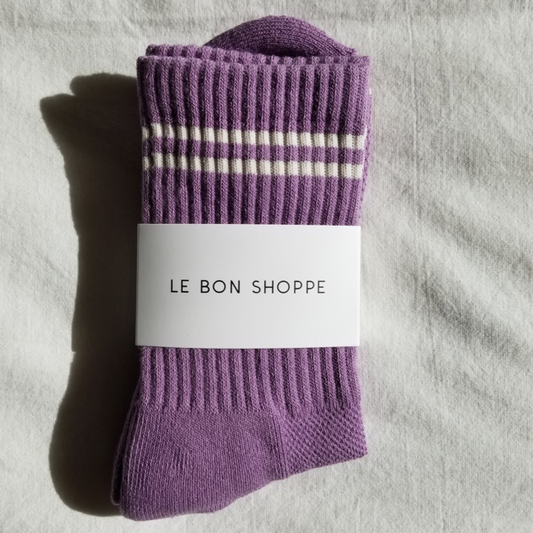A pair of grape purple ribbed crew socks with white stripes near the cuff, neatly folded and wrapped with a white Le Bon Shoppe label, placed on a white fabric background.
