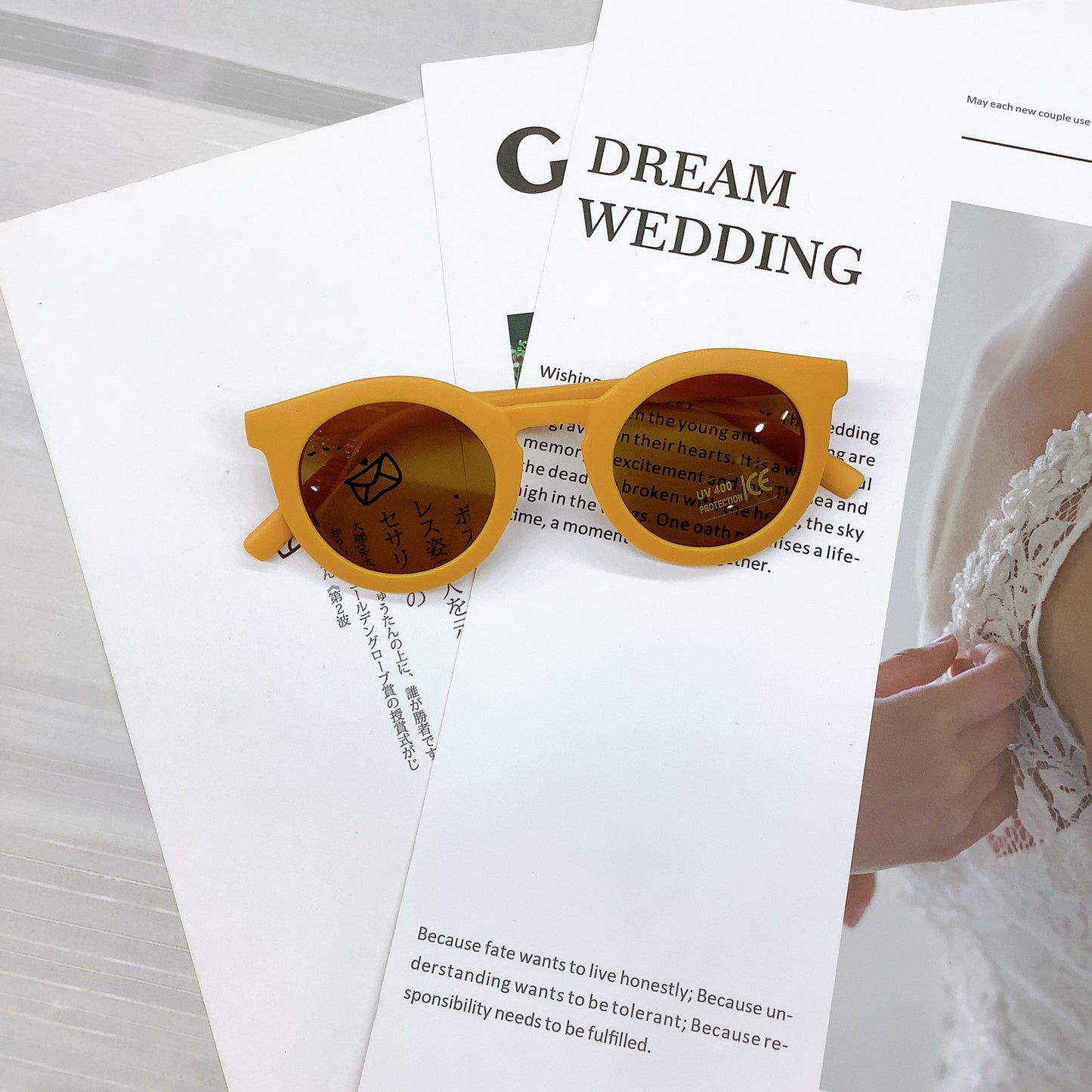 Kids' sunglasses in a warm marigold yellow colour with a round frame and UV400 protective lenses, casually placed on wedding-themed printed pages featuring text and a lace bridal detail in the background.