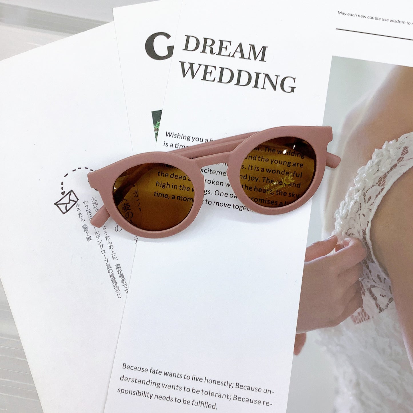 Kids' sunglasses in a soft mauve colour with a round frame and UV400 protective lenses, stylishly placed on wedding-themed printed pages with text and a bridal lace detail in the background.