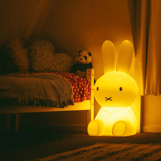 Mr Maria Miffy XL Lamp, 80 cm tall, iconic statement piece with six brightness settings, inspired by Dick Bruna's classic illustrations. It is lit up in a childs darkened  bedroom 