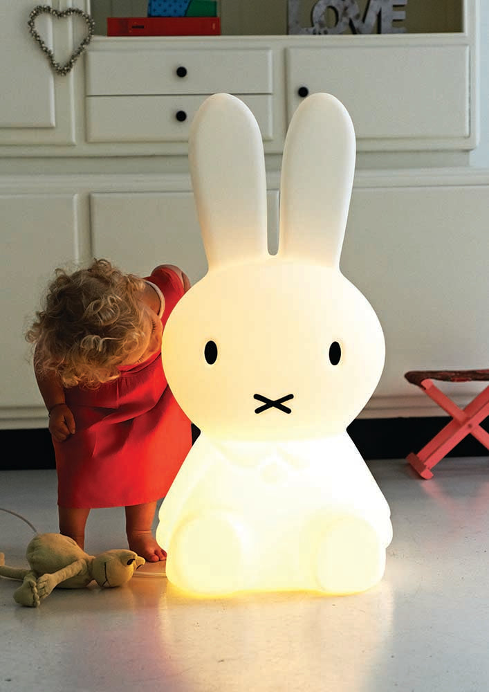 Mr Maria Miffy XL Lamp, 80 cm tall, iconic statement piece with six brightness settings, inspired by Dick Bruna's classic illustrations. It is lit up and small toddler in e red dress stands next to it for scale