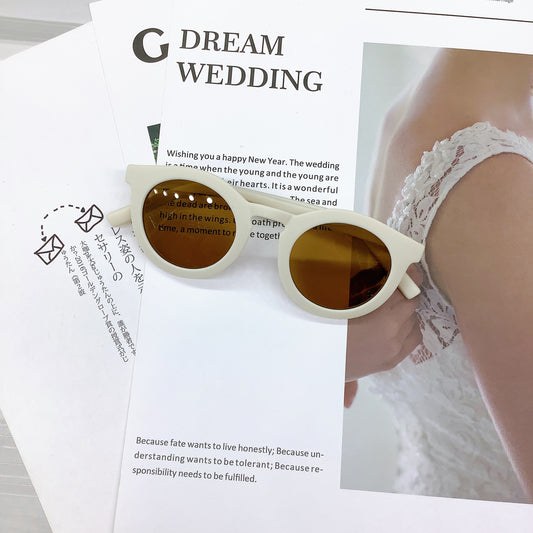 Kids' sunglasses in a milky white colour with a round frame and UV400 protective lenses, casually placed on wedding-themed printed pages featuring text and a lace bridal detail in the background.