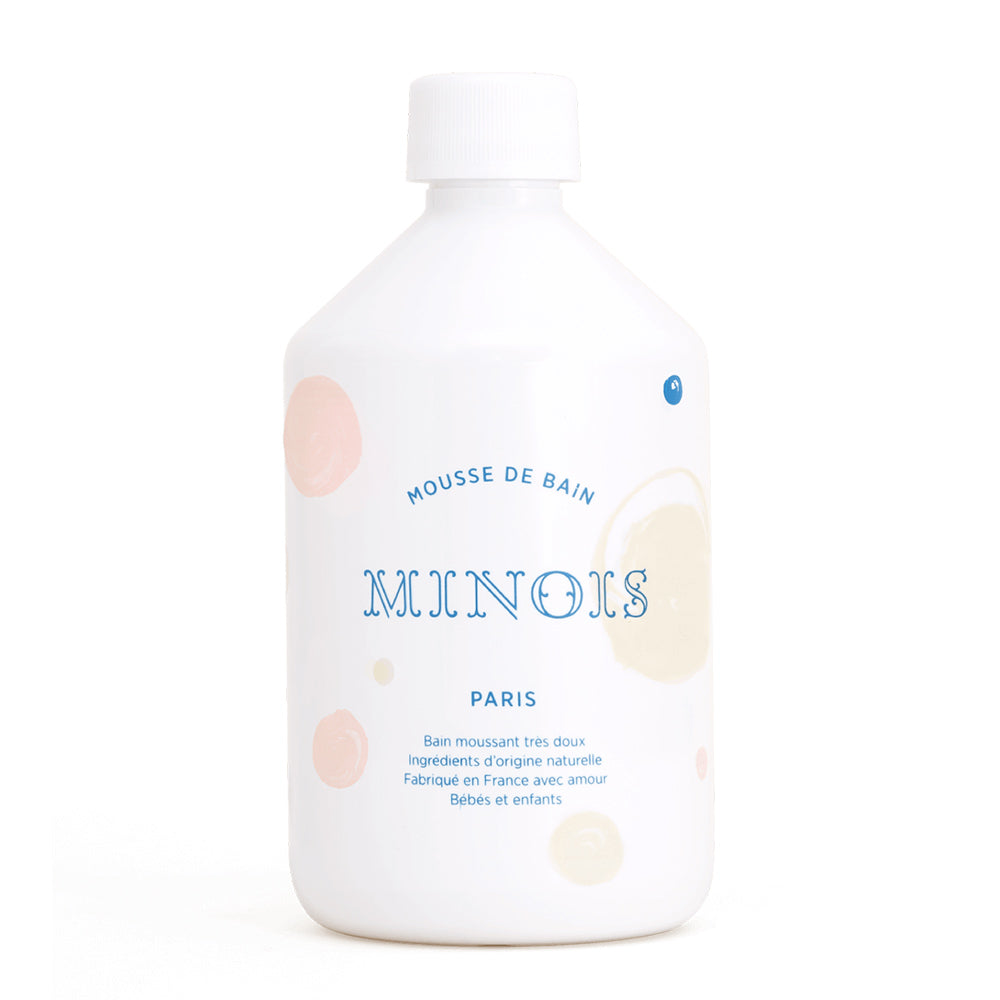 A white bottle of bubble bath, with a white screw on lid, that in blue says Mousse De Bain at the top which means bath foam, beneath that it says Minois, beneath that it says Paris. Beneath Paris there is smaller text in French which describes the ingredients of the product, where it was made and its suitability for children. The bottle itself is decorated with pink and beige spots. This is against a white background. 