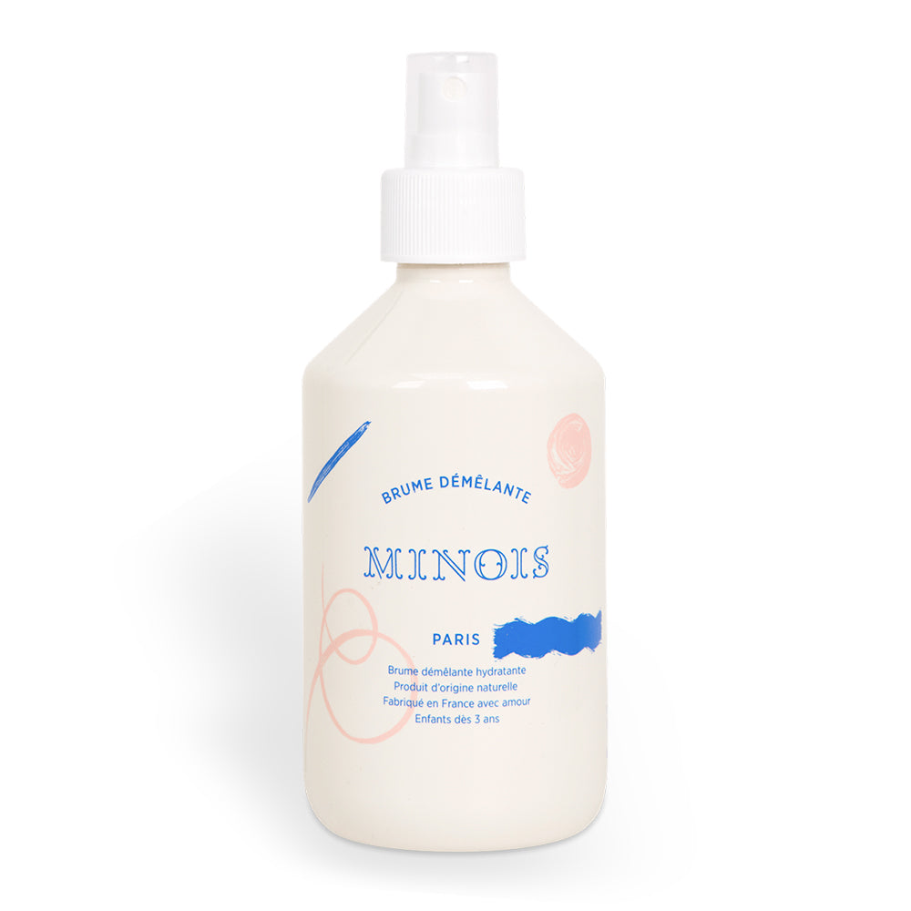 An off-white/cream bottle with a spray cap and lid sits in front of a white background. In blue it says Brume Demelante, meaning Detangling Mist and beneath that it says Minois, followed by Paris underneath that. Beneath Paris there is smaller text in French which describes the ingredients of the product, where it was made and its suitability for children. The bottle itself is decorated with pink spots and swirls and blue lines and patterns.