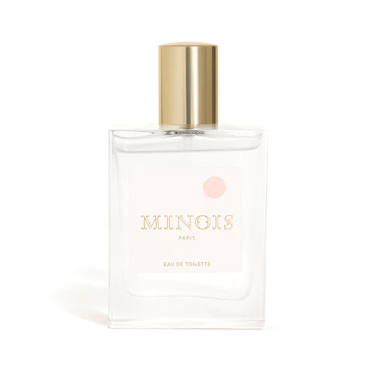 A clear square bottle of perfume with a gold lid sits in front of a white background. The bottle has a white label that says Minois in gold with a pink spot above it. Beneath Minois, also in gold it says Paris and beneath that, again in gold, it says Eau De Toilette. 