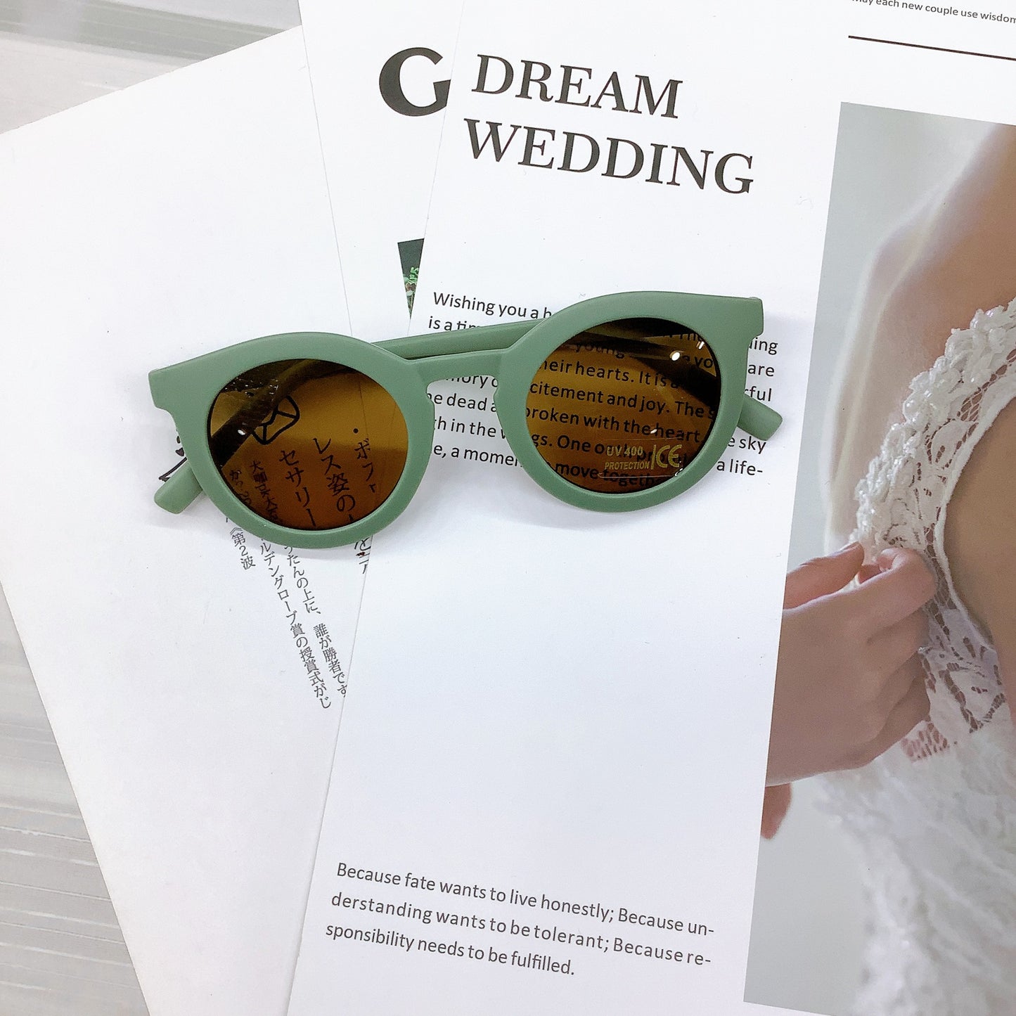 Kids' sunglasses in a stylish mint green colour with a round frame and UV400 protective lenses, casually placed on wedding-themed printed pages featuring text and a lace bridal detail in the background.