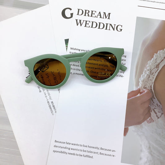 Kids' sunglasses in a stylish mint green colour with a round frame and UV400 protective lenses, casually placed on wedding-themed printed pages featuring text and a lace bridal detail in the background.