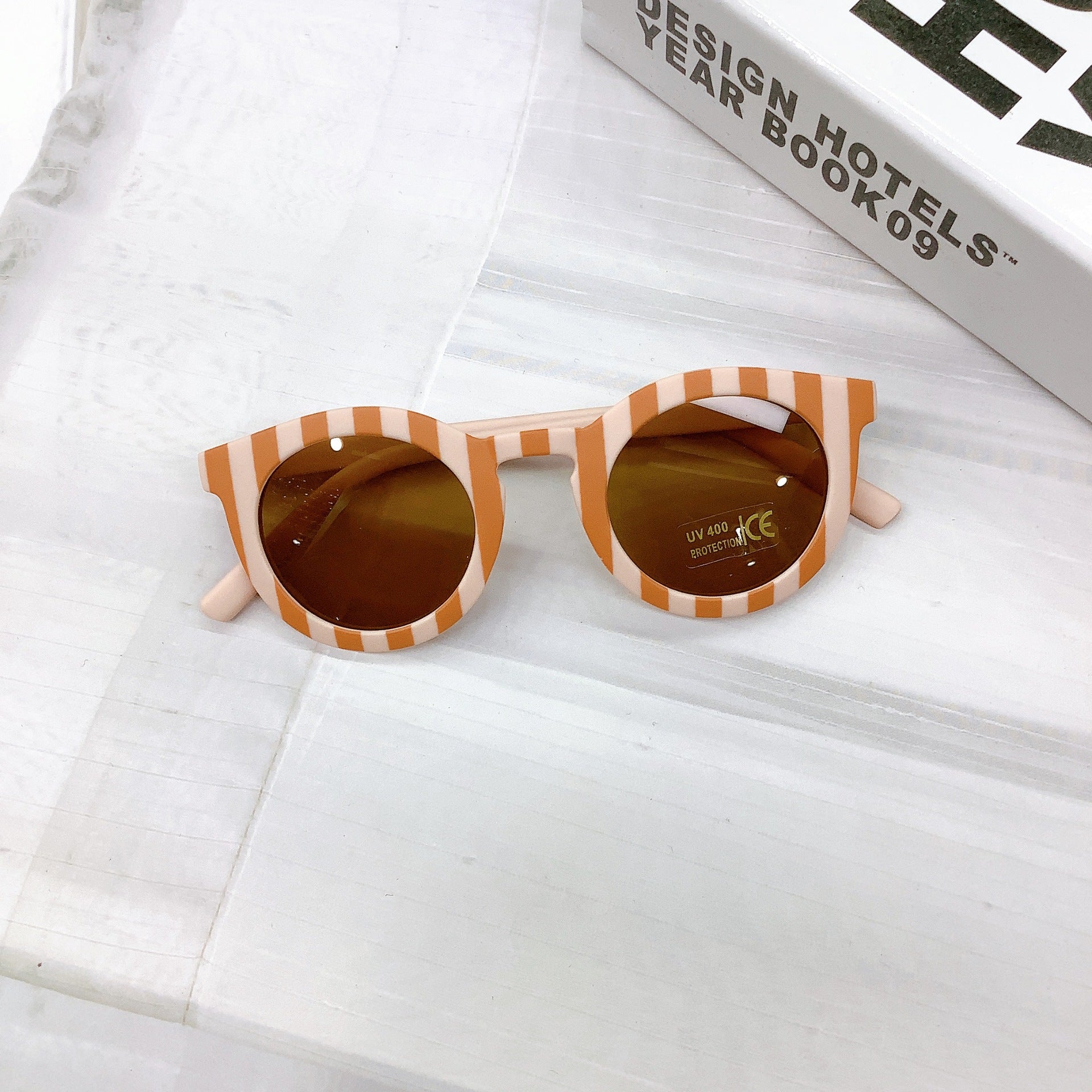 Kids' sunglasses with a bold mustard and cloud pink stripe frame, round UV400 protective lenses, and a playful retro design, displayed on a white surface next to a design book
