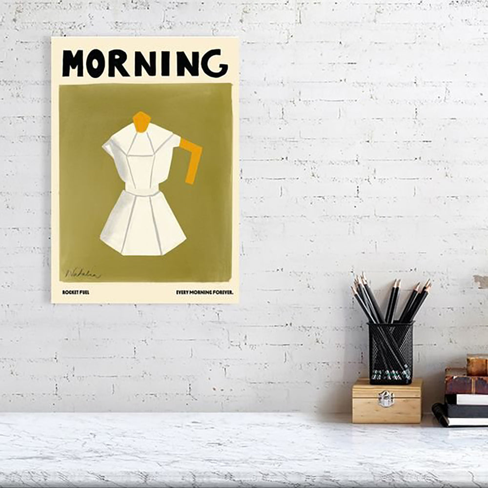 A Natalia Bagniewska Morning Print in a room setting. In the print, the background is bright olive green. The jug is white with grey edges. The lid and holder of the jug is ochre yellow in colour. The print is framed and hung on a white wall. The table is white with a wooden box on top. The pencils and the pencil stand are black. 
