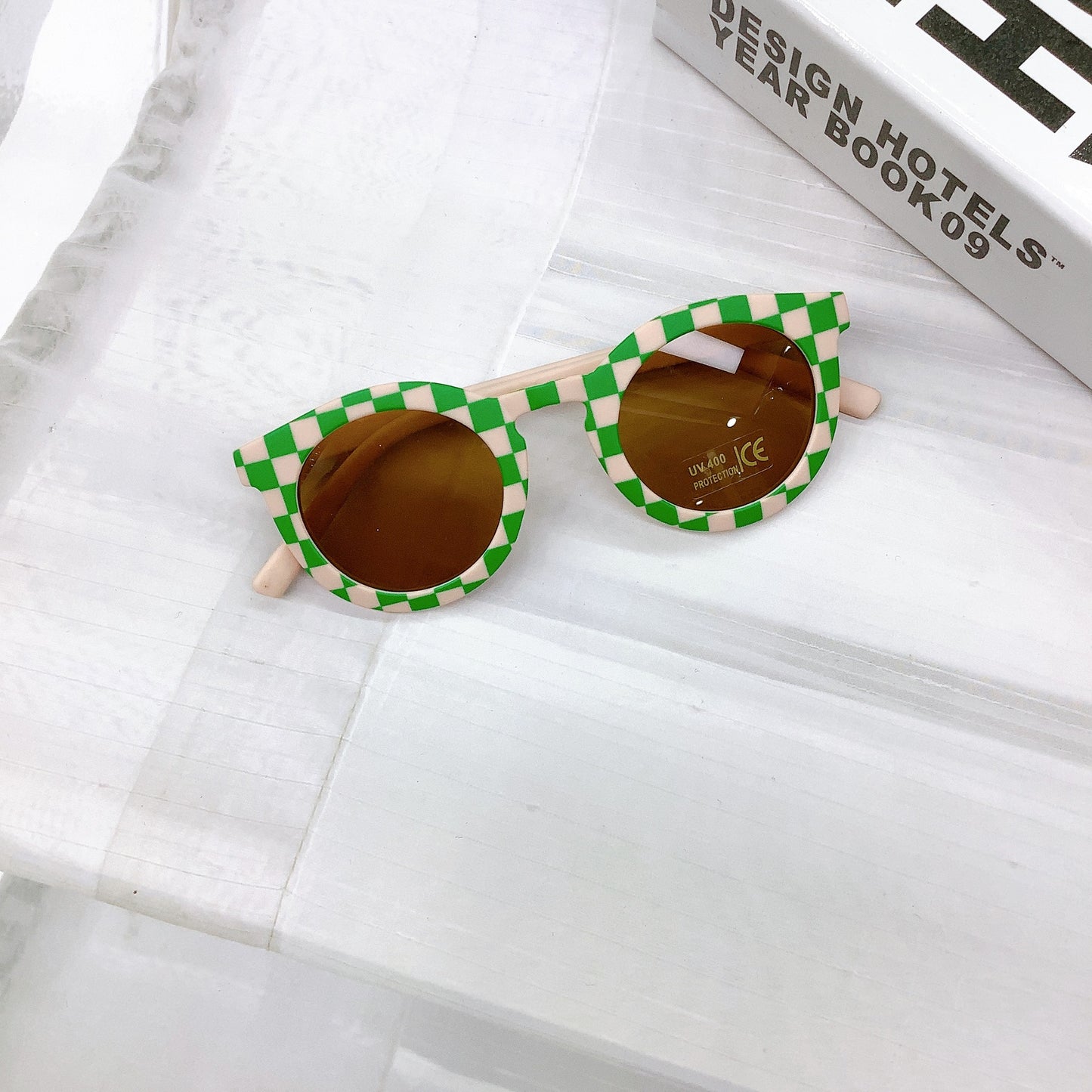 Kids' sunglasses with a bold peach and green check frame, round UV400 protective lenses, and a playful retro design, displayed on a white surface next to a design book