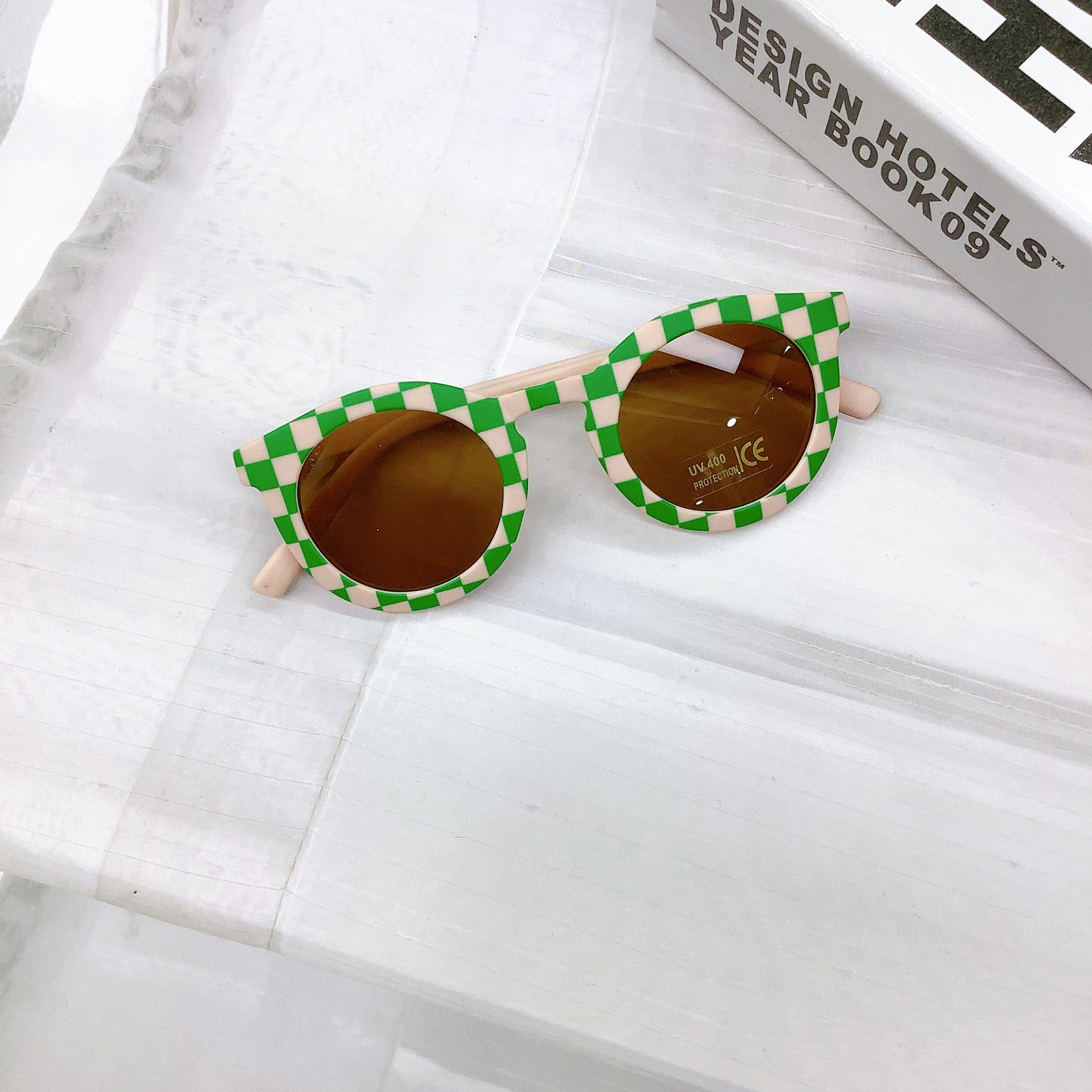 Kids' sunglasses with a bold peach and green check frame, round UV400 protective lenses, and a playful retro design, displayed on a white surface next to a design book