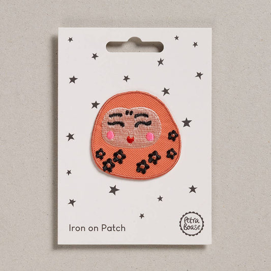 an embroidered daruma doll that is peachy orange in colour with black flowers. The doll’s face is shiny and light pink with pink blush and red lips. The background is white with black stars. 