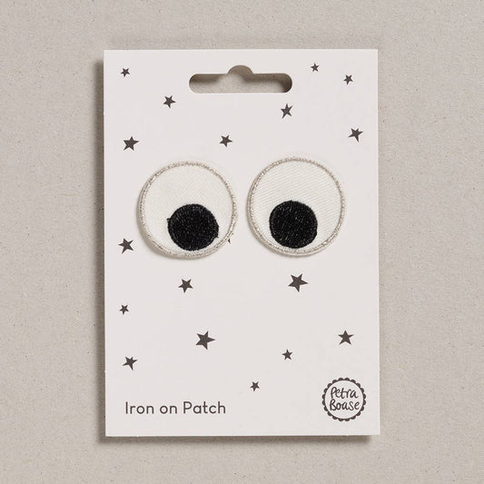 an embroidered googly eyes patch mainly white with black pupils and silver trim. The background is white with black stars. 