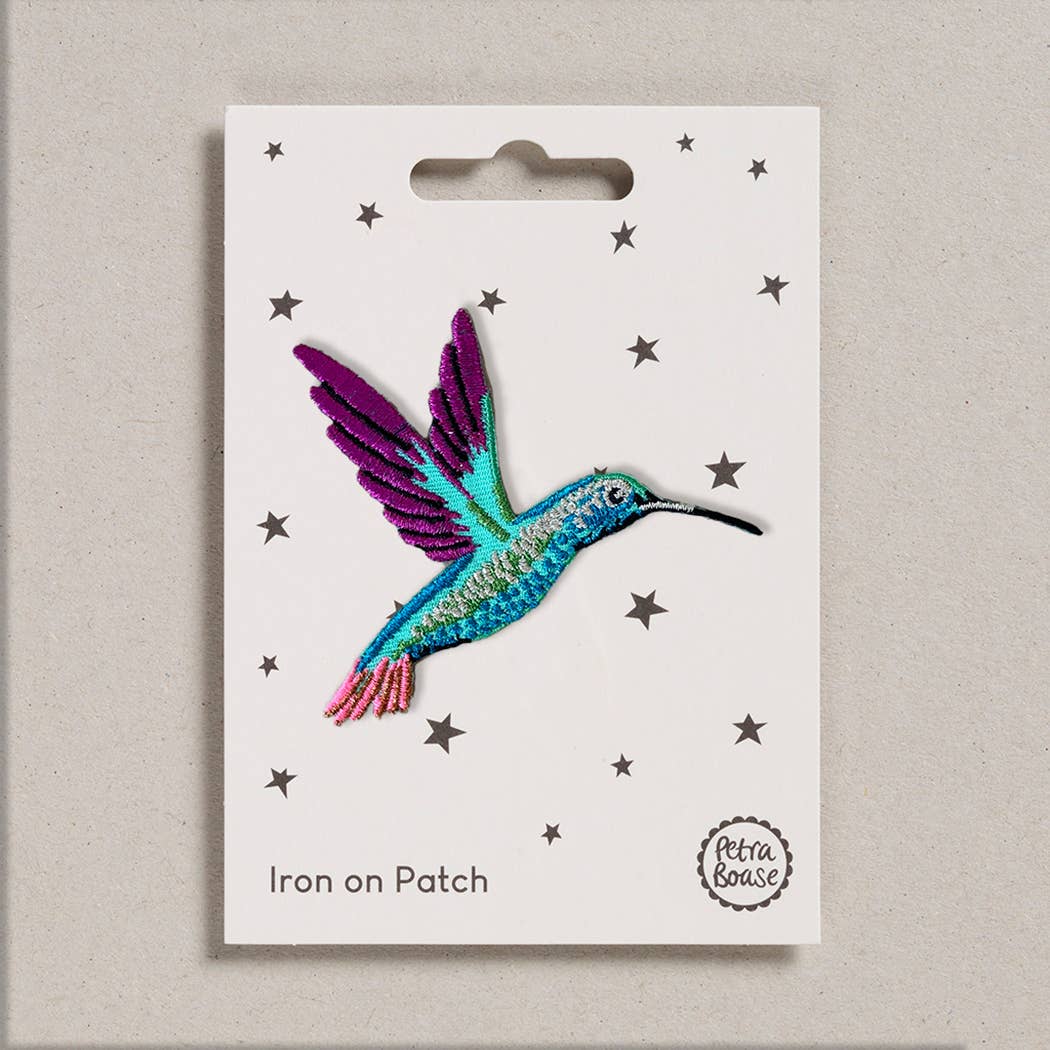 An embroidered iron-on patch shaped like a vibrant hummingbird in flight, featuring shimmering turquoise, purple, and pink stitching with intricate wing and beak detailing. Packaged on a cream backing card with black stars and Petra Boase branding.
