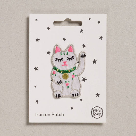 an embroidered white Lucky cat. The eyes are black. The ears, nose and mouth are pink. The collar around its neck is green and golden with a golden pendant. The cat has pink flowers and green leaves on its body. The background is white with black stars. 