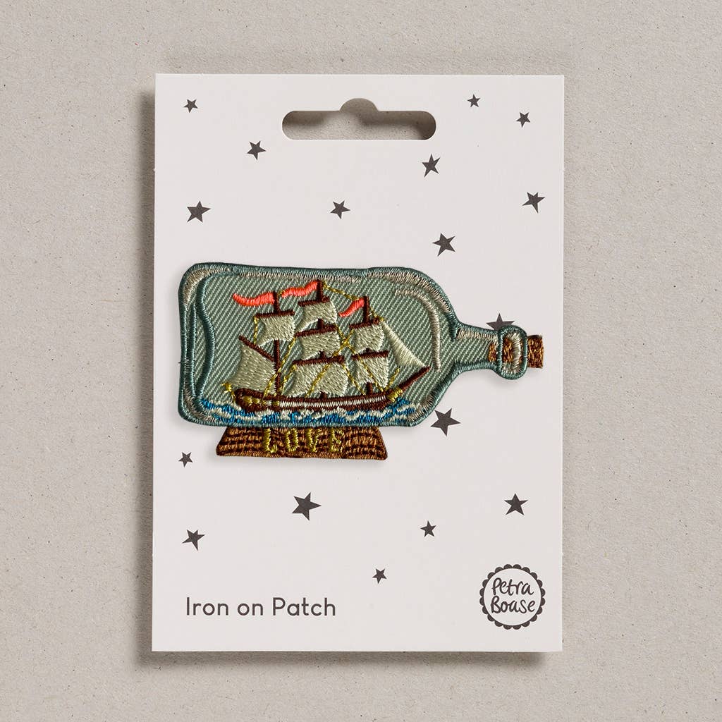 An embroidered iron-on patch featuring a classic tall ship with red sails inside a glass bottle, resting on a wooden stand with "Hope" stitched in gold. Packaged on a cream backing card with black stars and Petra Boase branding.