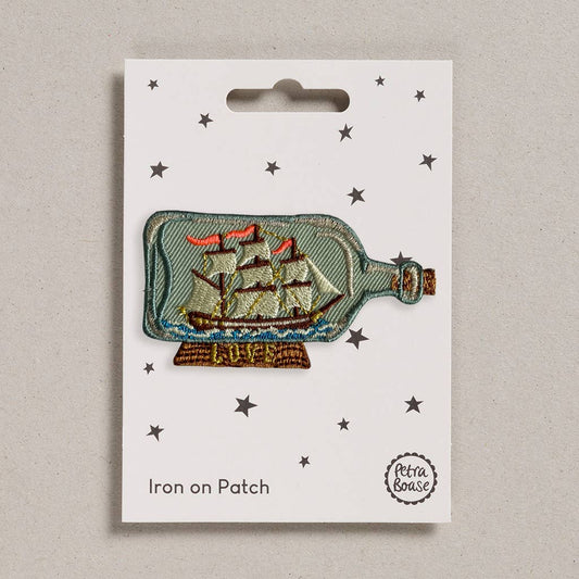 An embroidered iron-on patch featuring a classic tall ship with red sails inside a glass bottle, resting on a wooden stand with "Hope" stitched in gold. Packaged on a cream backing card with black stars and Petra Boase branding.