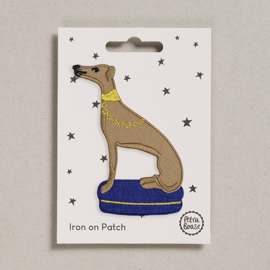 an embroidered whippet who is light brown is colour. His nose and eyes are black. He has a golden collar around his neck with a golden leash. He sits on a blue cushion with a golden line. The background is white with black stars. 