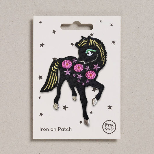 an embroidered black pony with golden tail and mane. She has silver feet, green eyelashes and silver eyes. Her body is adorned with pink stars and flowers. The background is white with black stars