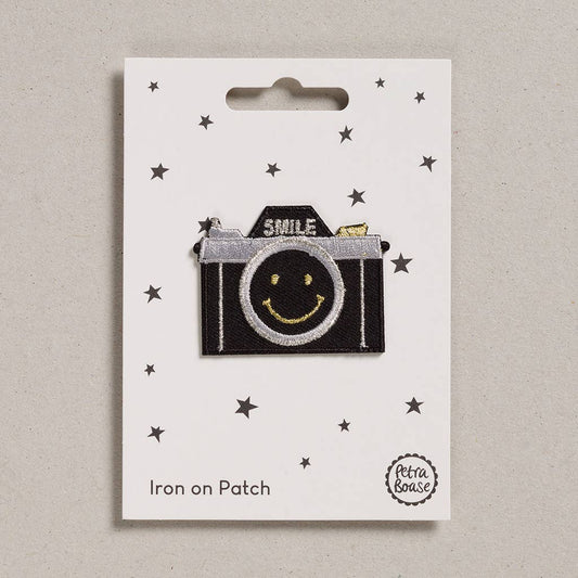 an embroidered patch of a black film camera with a smiley face on the lens with gold stitching and the word smile where the flash is in silver thread