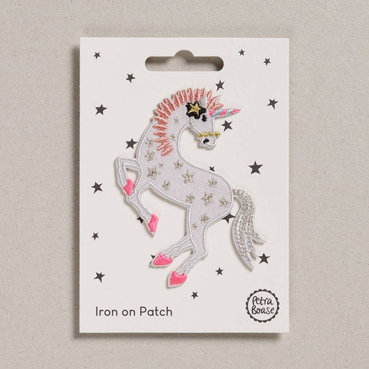 an embroidered unicorn. The unicorn is white in colour with silver tail and pink mane. Her feet are pink and her body is adorned with silver stars. She has a black and pink star near her ear. Her horn is pink and blue. The background is white with black stars