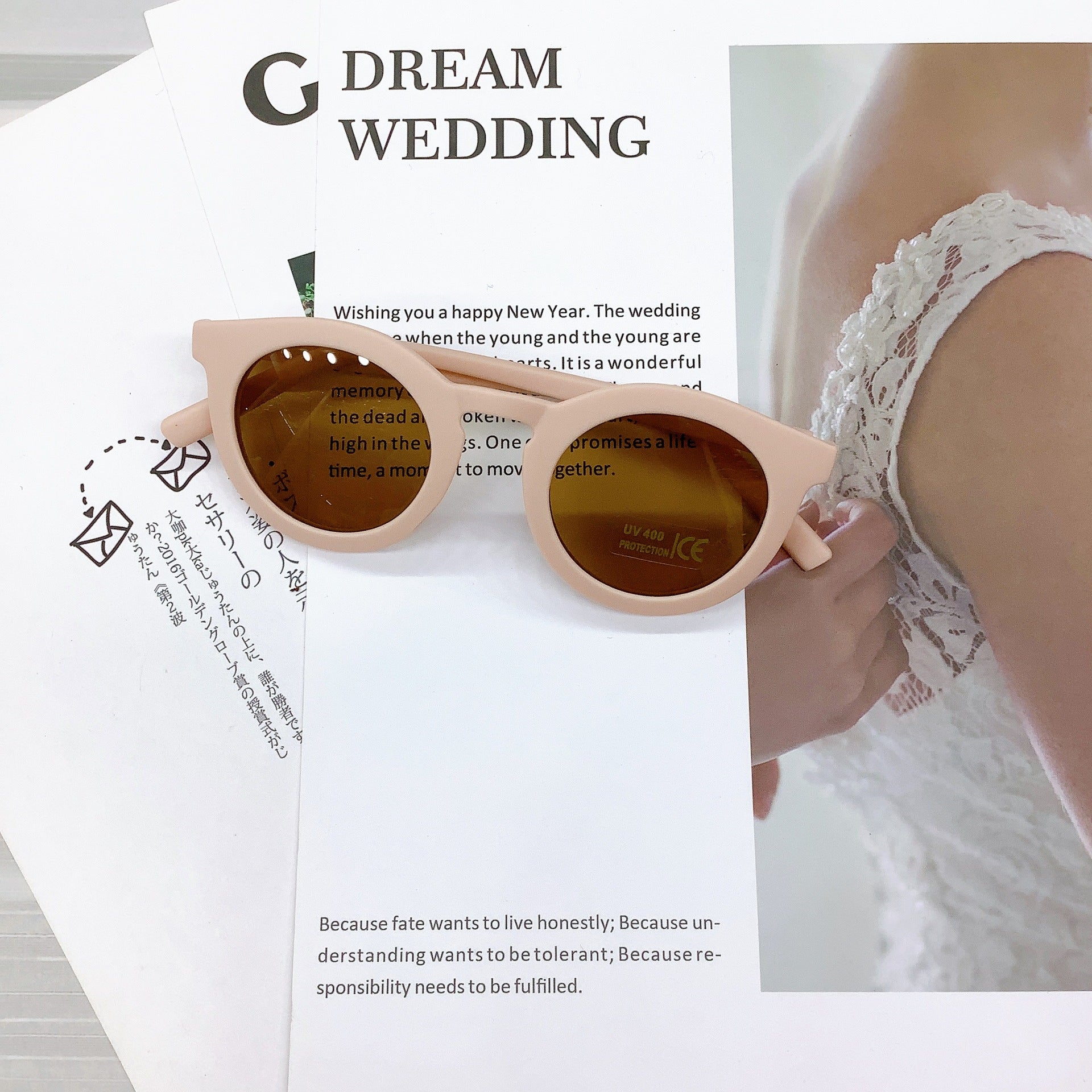Kids' sunglasses in a soft cloud pink colour with a round frame and UV400 protective lenses, casually placed on wedding-themed printed pages featuring text and a lace bridal detail in the background.