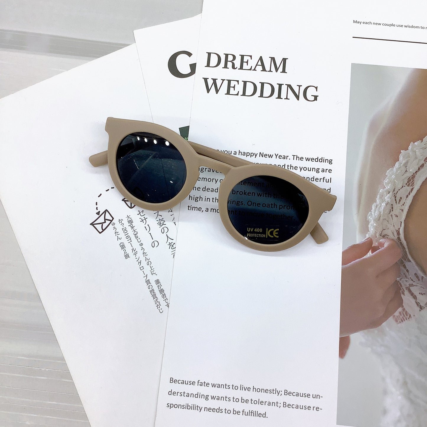 Kids' sunglasses in a stylish taupe colour with a round frame and UV400 protective lenses, casually placed on wedding-themed printed pages featuring text and a lace bridal detail in the background.