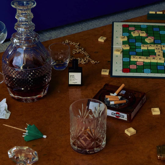 An image for a table which on it has a crystallised glass with brown liquid in it, a creen cocktail stick, a dirty ashtray, a small shot size clear glass, a scrabble board with letters scattered across the table, a liqour decanter with brown liquid inside and a bottle of the gueule de bois perfume. 